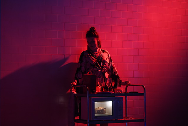 A person in a robe stands behind a microwave. Red hue in the lighting.