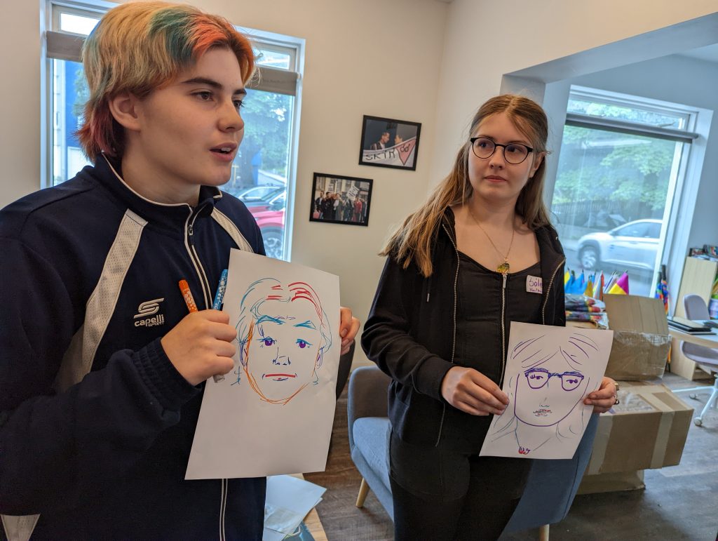Two young people indoors, one with black glasses and long hair and another with short, multi-colored hair, hold up doodled portrait of themselves.