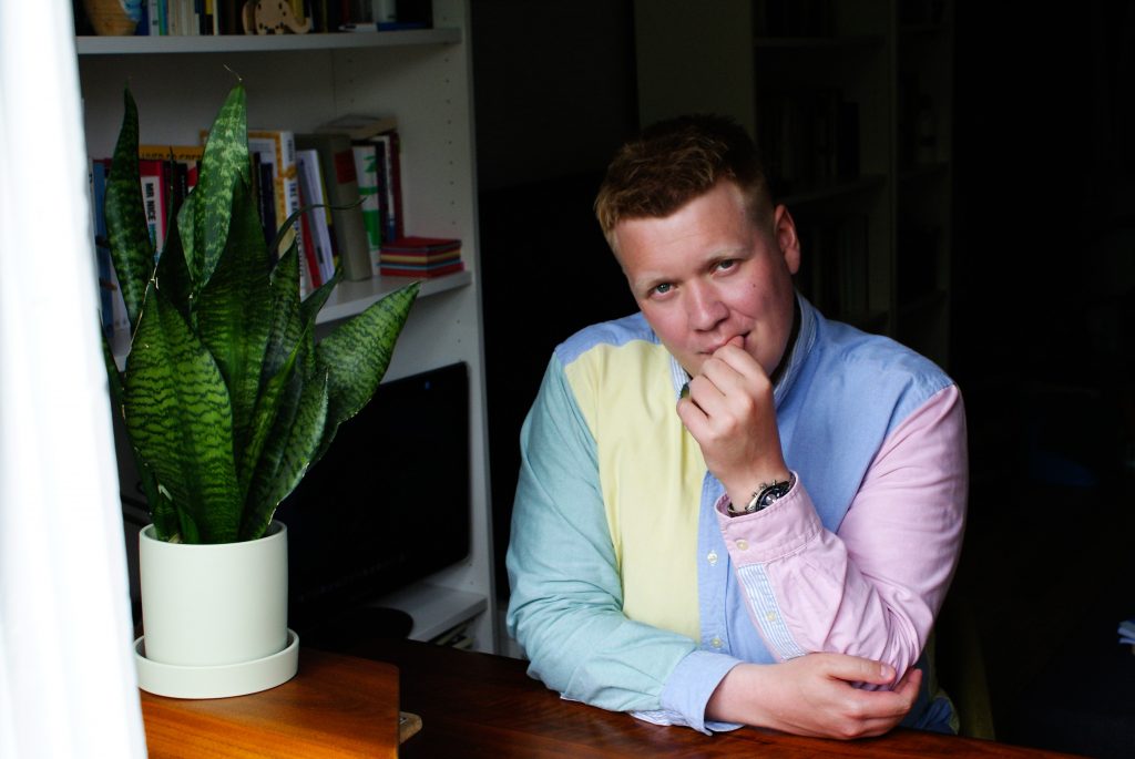 A man in a pastel colored shirt bites his nails. A plant sits on the table next to him, in front of a bookcase.