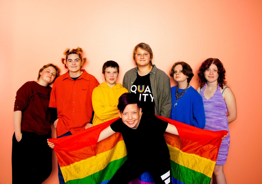 A group of young people, posing in front of a studio background. THey are holding a pride flag. 