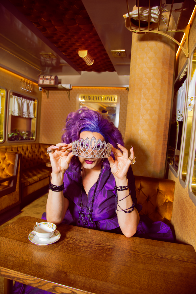 A person with purple hair in purple clothes has a tiara in front of their eyes