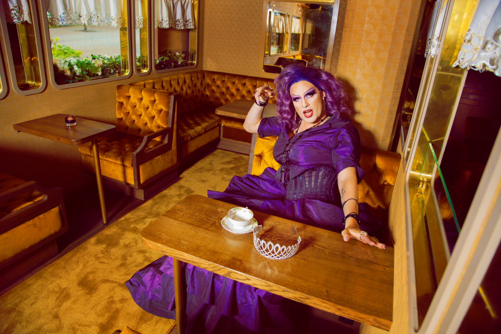 A drag queen in purple with a purple wig sits by a table with a cup of coffee and a grown in front of her.