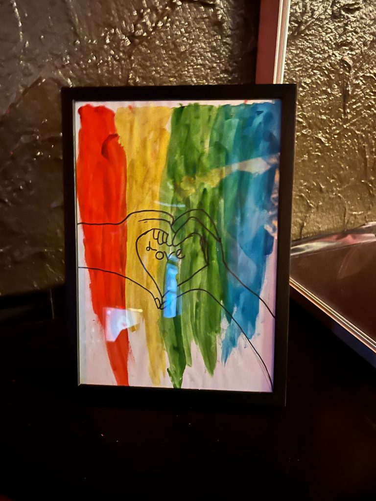 A painting with two hands forming a heart in front of a rainbow
