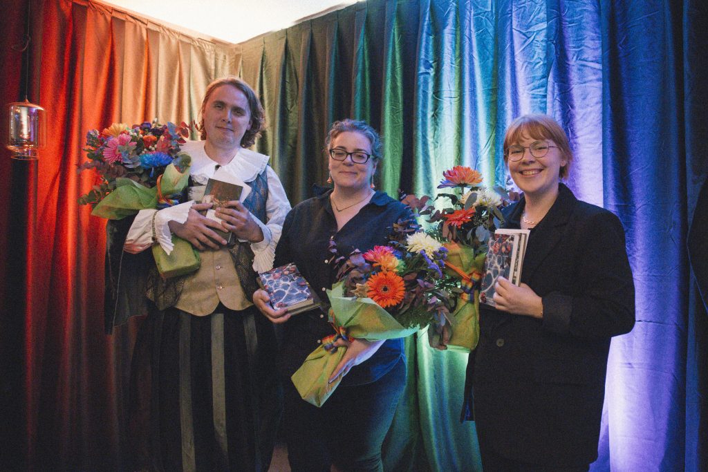 Three people with flower bouquets and prizes, in front of a rainbow tapestry