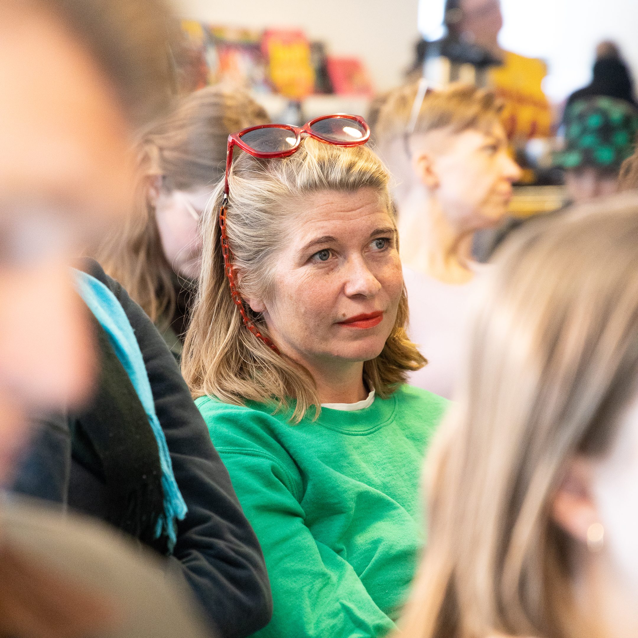 A blonde woman in a green sweater sits in a crowd of people. She has red sunglasses on her head and a red lipstick.